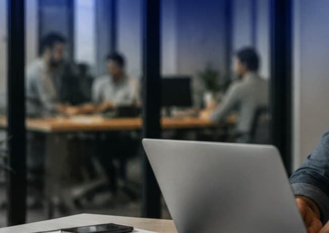 Escritório com equipe em reunião ao fundo e notebook em primeiro plano